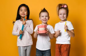 A boy holding a model of teeth with two girls holding large toothbrushes in front of an orange background